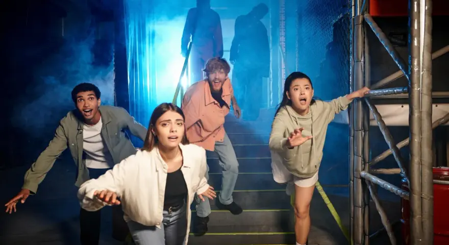 Grupa przerażonych ludzi ucieka przed postaciami w dymie. To wygląda jak prawdziwy escape room, gdzie trzeba rozwiązać zagadki, by przeżyć.