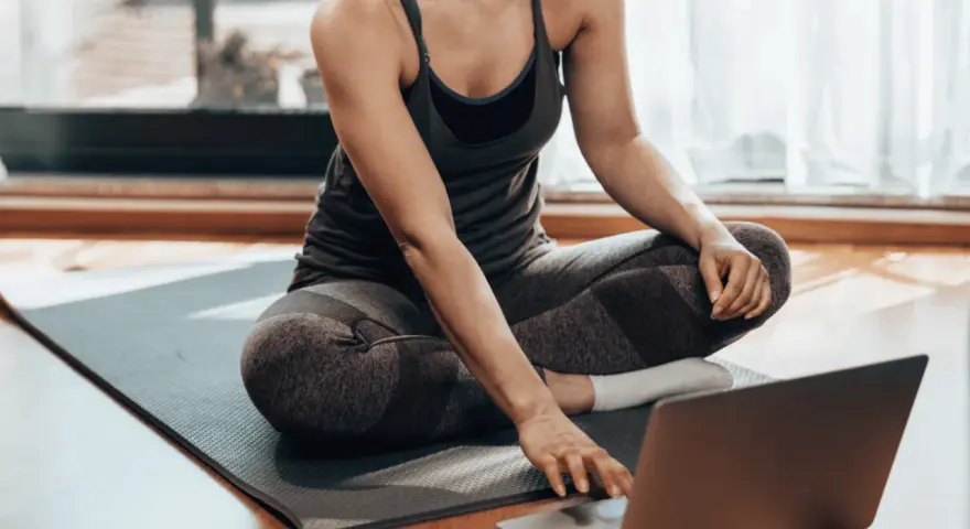 Kobieta w stroju sportowym siedzi na macie, korzystając z laptopa. Czy można ćwiczyć codziennie? Tak, jeśli masz dostęp do inspiracji online!
