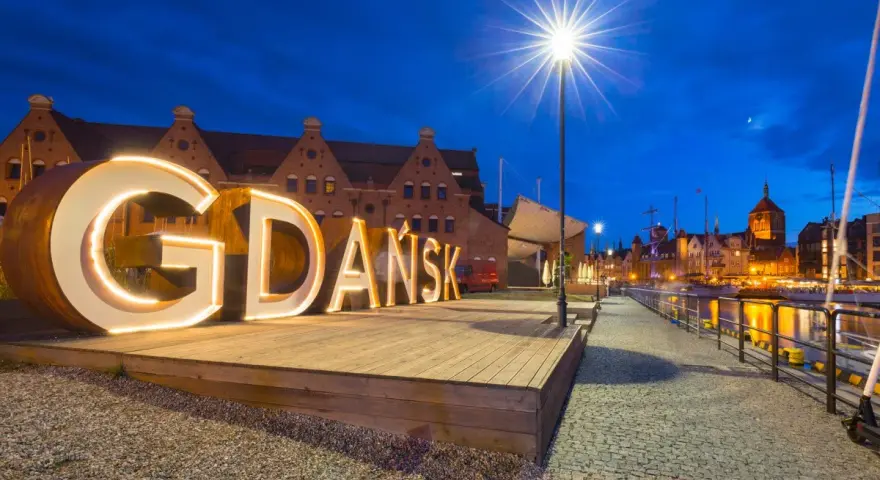Podświetlany napis "GDAŃSK" na tle nocnej panoramy miasta, zapraszający do odkrywania atrakcji Trójmiasta.