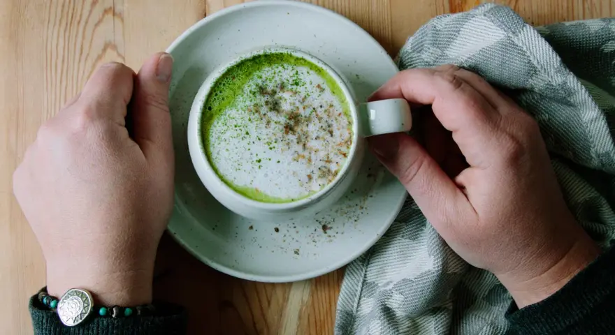 Händerna håller en kopp med en krämig matcha latte, toppad med skum och lite kryddor. En mysig stund med en grön dryck.