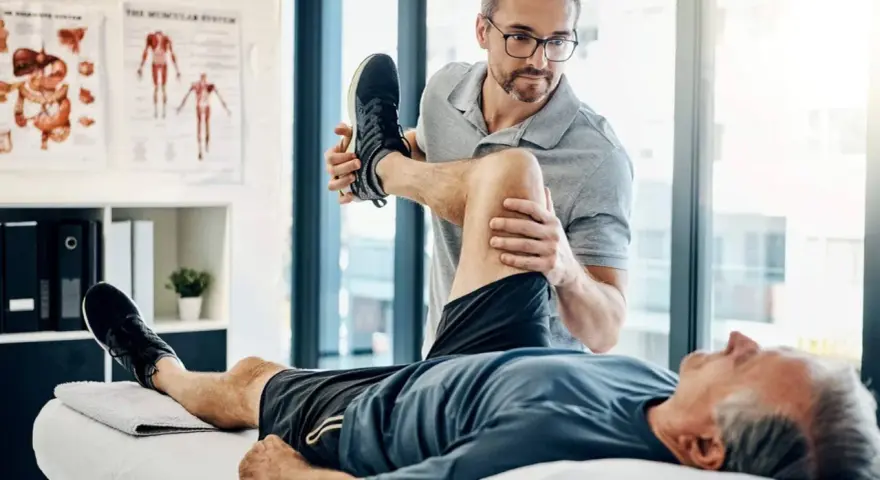 Ein Physiotherapeut dehnt das Bein eines Patienten. Der Patient liegt auf einer Liege, während der Therapeut sein Knie beugt. Wann Physiotherapie? Hier wird geholfen.