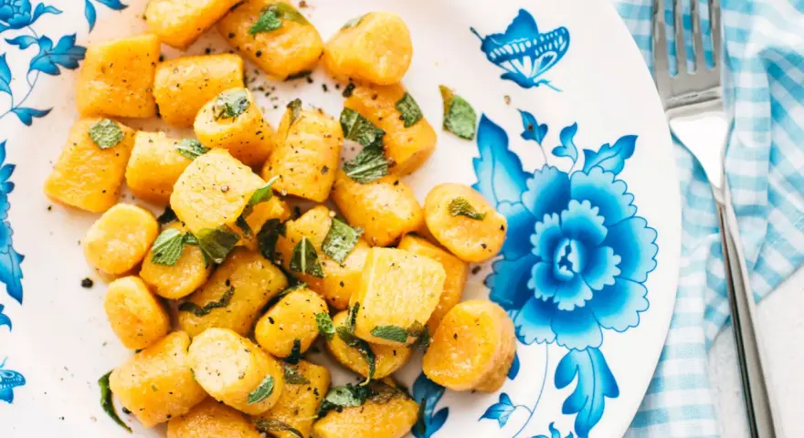 Gnocchi di zucca dorati, conditi con erbe aromatiche e pepe nero, su un piatto decorato con fiori blu.