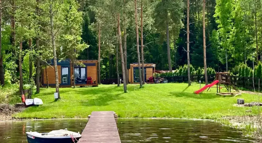 Drewniany pomost nad jeziorem Bardze, z łódką przycumowaną do brzegu. W tle domki letniskowe i plac zabaw.