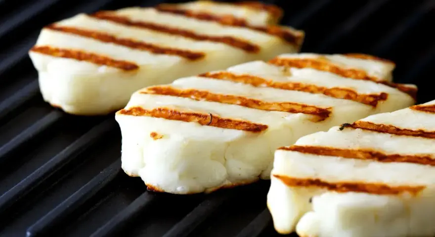 Trzy kawałki halloumi z grilla, z apetycznymi paskami, leżą na czarnej grillowej płycie.