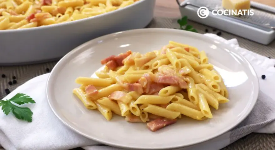 Plato de macarrones a la carbonara cremosos con trozos de panceta. En el fondo, un bol con más pasta y un rallador de queso.