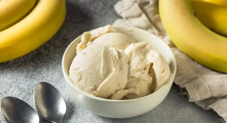 Un cremoso helado de plátano en un tazón blanco, con plátanos amarillos frescos al lado.