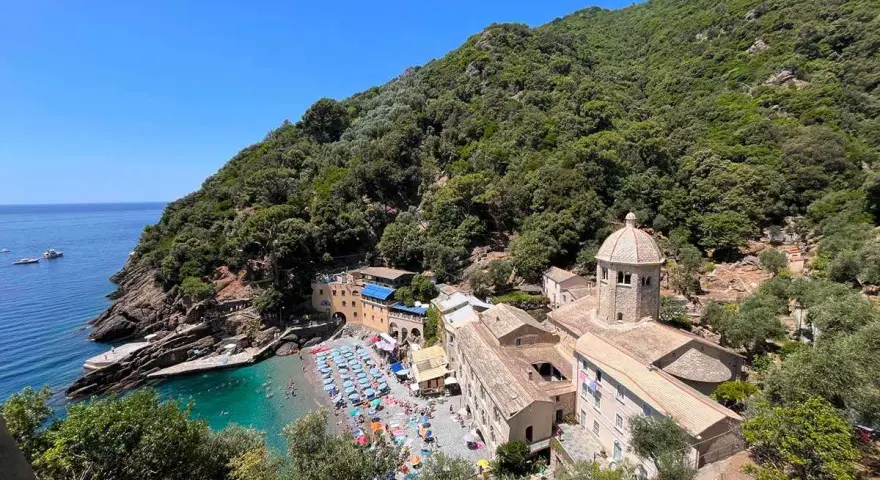 Baia con spiaggia affollata e un antico monastero, un luogo ideale per i cammini in Liguria, con mare azzurro e vegetazione rigogliosa.