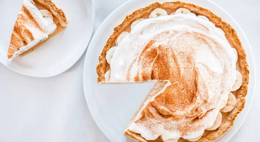 Une part de tarte banoffee traditionnel, garnie de crème fouettée et de cannelle, sur une assiette blanche.