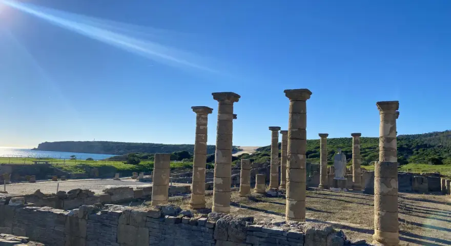 Sunlight streams over ancient roman ruins in Spain, illuminating weathered stone columns and walls against a bright blue sky and distant coastline.