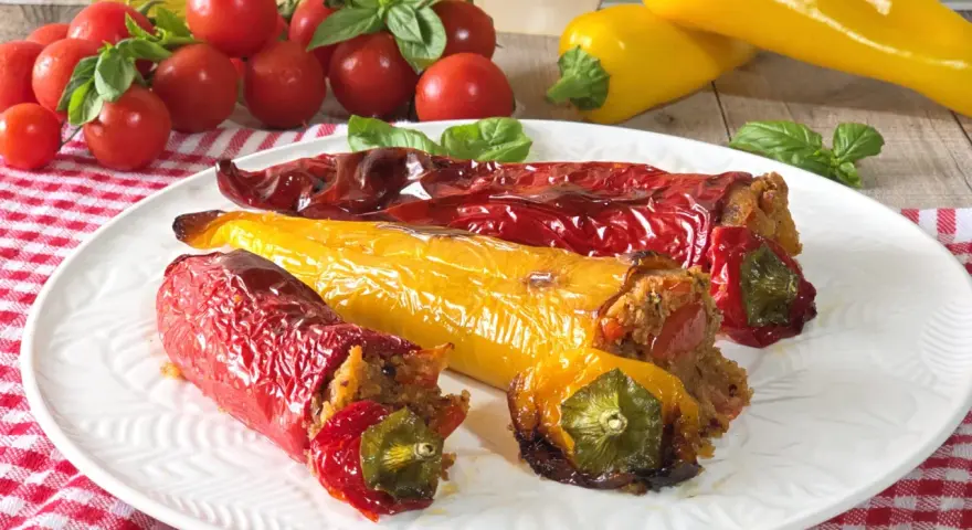 Peperoni cornetti ripieni, rossi e gialli, cotti al forno, su un piatto bianco con pomodorini e basilico.