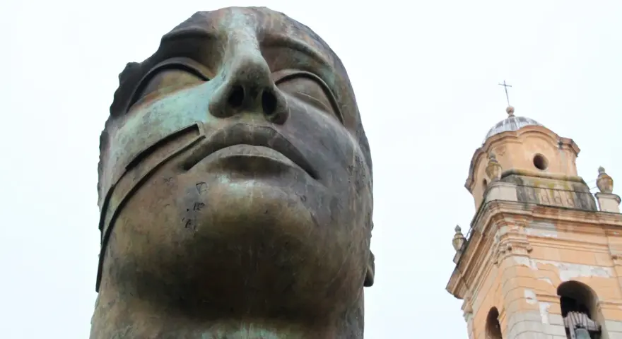 Brązowa, monumentalna rzeźba głowy z widocznym fragmentem dzwonnicy kościelnej w tle.