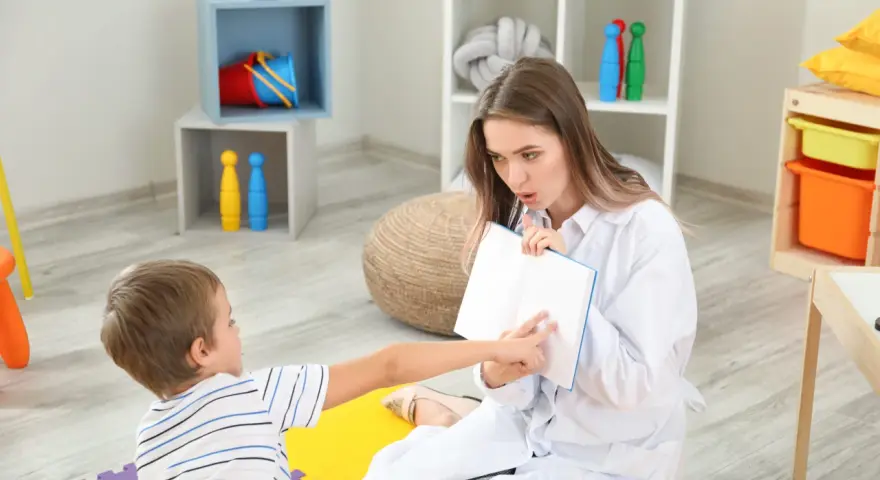Terapeuta pracuje z dzieckiem, pomagając w rozwoju mowy. Chłopiec wskazuje na książkę, a kobieta uważnie go słucha.