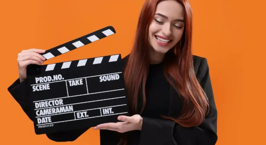 Uśmiechnięta kobieta z rudymi włosami trzyma klaps filmowy. Zastanawiasz się, ile kosztuje hollywood smile w Polsce?