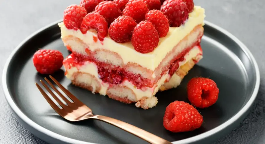 Pyszne tiramisu malinowe z malinami na wierzchu i na talerzu, obok złoty widelec.