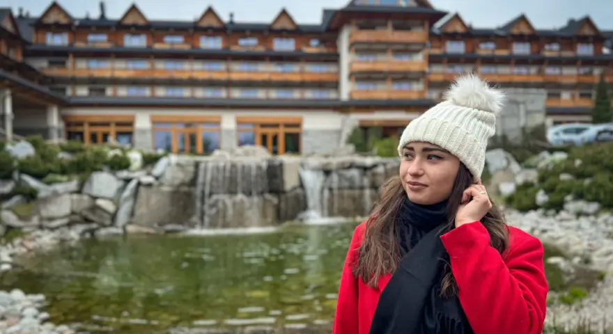 Kobieta w czerwonym płaszczu i białej czapce przed hotelem Bania. W tle wodospad i staw. Wiele pozytywnych hotel Bania opinie potwierdza ten widok.