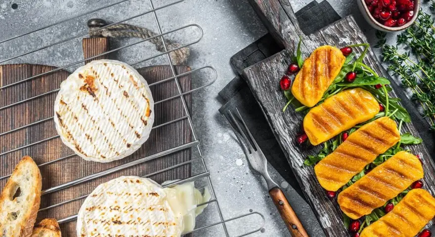 Dwa okrągłe serki na grilla, jeden lekko roztopiony, obok pieczywo. Na desce grillowane kawałki dyni z granatem i rukolą.