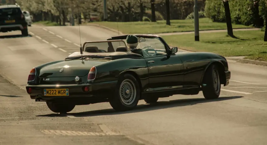Auto d'epoca verde scuro in movimento, con conducente con cappello. La targa suggerisce che siamo in Inghilterra, dove si guida a destra.
