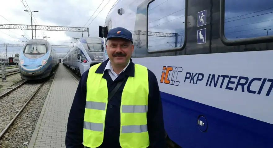 Uśmiechnięty rewident taboru kolejowego w kamizelce odblaskowej stoi obok pociągu PKP Intercity.