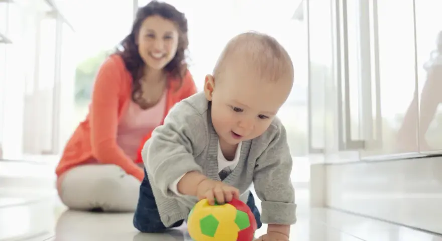 Maluch ćwiczy motorykę dużą, raczkując po podłodze i bawiąc się kolorową piłeczką. Uśmiechnięta mama obserwuje jego postępy.