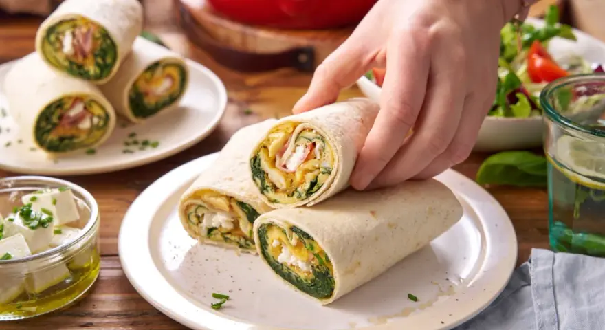 Ręka sięga po tortillę ze szpinakiem, jajecznicą i serem feta. Na talerzu leżą kolejne zawiniątka, obok miseczka z fetą i sałatka.