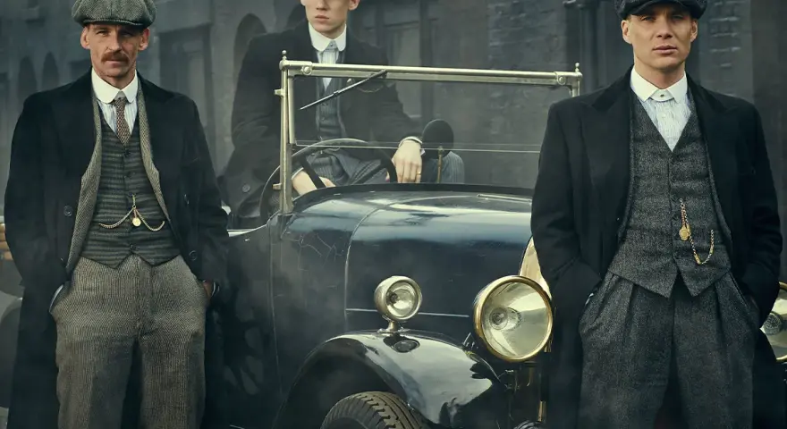 Tre uomini in abiti eleganti, stile Peaky Blinders, posano accanto a un'auto d'epoca.