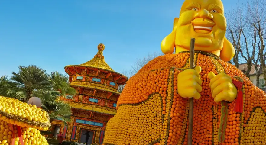 Escultura gigante de Buda hecha de naranjas y limones, parte del menton lemon festival, con una pagoda de frutas al fondo.
