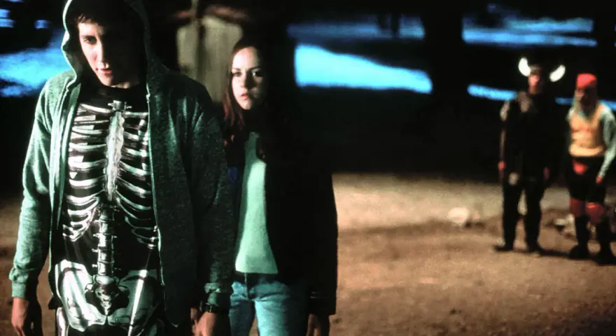 Donnie Darko w bluzie z kapturem i koszulce z nadrukiem szkieletu, obok dziewczyny. W tle dwie postacie w kostiumach.