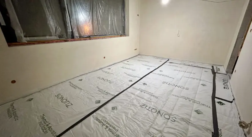 W trakcie prac remontowych w bloku, podłoga pokryta jest materiałem do wyciszenia. Widać okno zabezpieczone folią i sufit z widocznymi instalacjami.