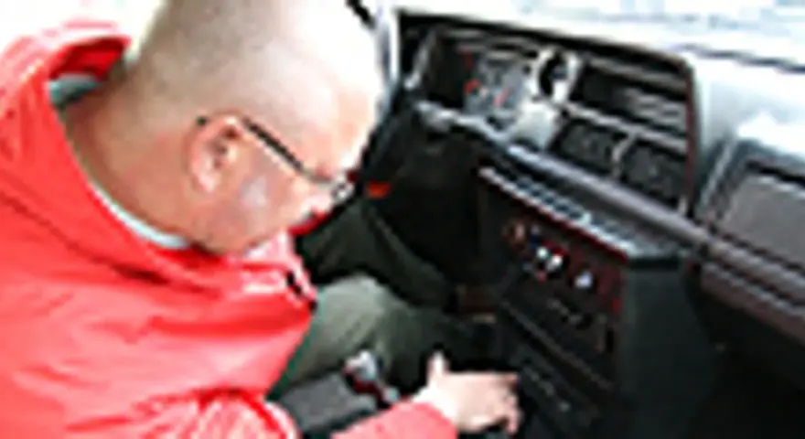 Mężczyzna w czerwonej kurtce pochyla się nad deską rozdzielczą, szukając rozwiązania, **co zrobić żeby radio nie szumiało**.