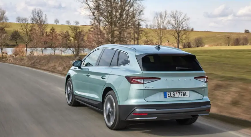 Jasnozielony Skoda Enyaq jedzie po wiejskiej drodze, otoczony wiosennym krajobrazem z drzewami i zielonymi polami.