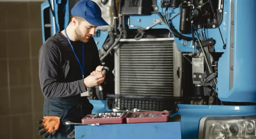 Mechanik w niebieskiej czapce i kombinezonie naprawia ciężarówkę, trzymając narzędzie.