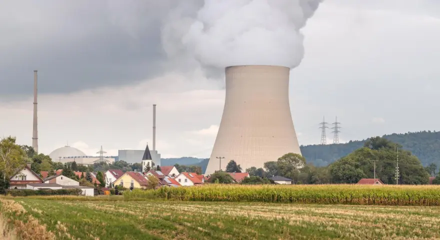 Dampf steigt aus einem Kühlturm eines Atomkraftwerks auf, daneben ein Dorf und ein Maisfeld.