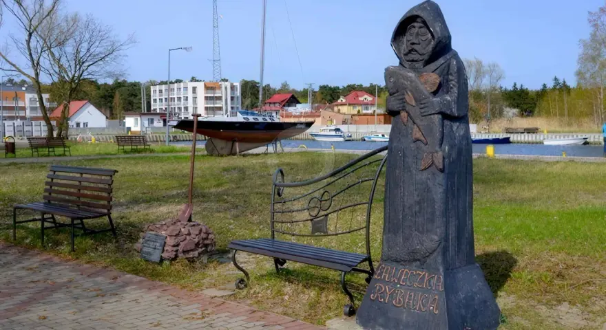 Drewniany pomnik rybaka trzymającego rybę, z napisem "Ławeczka Rybaka". W tle port z łodziami i budynkami.