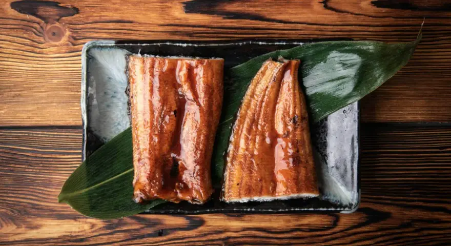 Dwa kawałki glazurowanego węgorza do sushi, podane na liściu na ciemnej zastawie, na drewnianym tle.