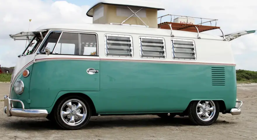 En turkos och vit volkswagen folkabuss med taklucka och takräcke står på en sandstrand.