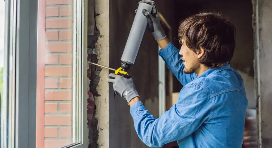 Mann mit Handschuhen und Schaumpistole dichtet den Rahmen für den Fenstertausch ab.