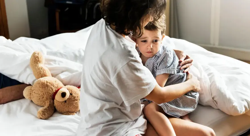 Mutter tröstet ihr Kind, das Anzeichen einer Panikattacke bei Kindern zeigt. Ein Teddybär liegt daneben.
