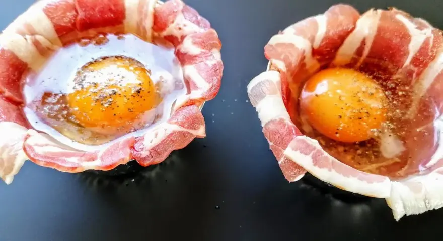 Due uova crude con tuorlo brillante, avvolte in strisce di bacon croccante, pronte per essere cotte.