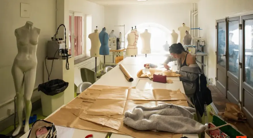 Dans un atelier d'une école d'arts, une jeune femme coupe du tissu sur une grande table. Des mannequins et du matériel de couture sont visibles.