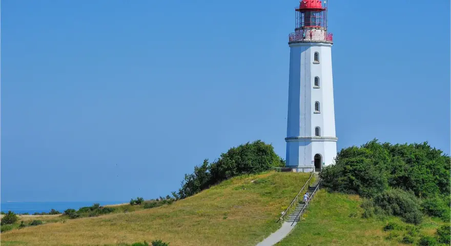 Der Leuchtturm Dornbusch ragt auf einer grünen Anhöhe empor, mit dem blauen Meer im Hintergrund. Eine Treppe führt zum Eingang.