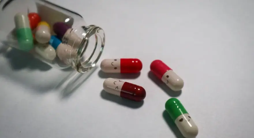 Kolorowe kapsułki, w tym leki psychotropowe, wysypują się ze szklanej buteleczki. Niektóre mają narysowane uśmiechnięte buźki.