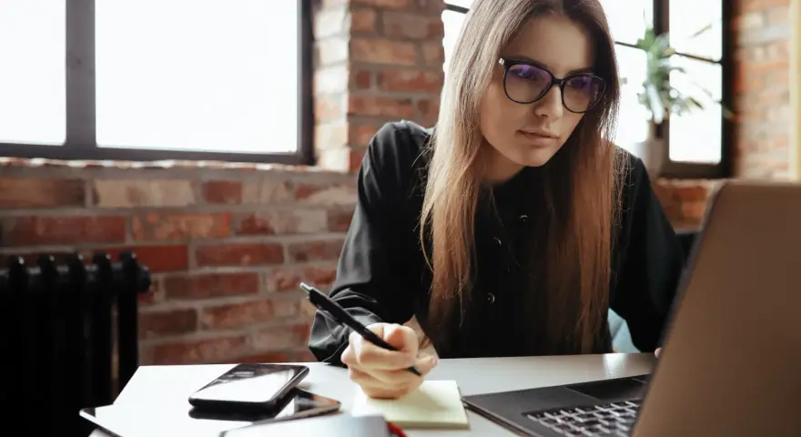 Młoda kobieta w okularach pisze notatki, przygotowując dodatek motywacyjny do CV. Na biurku laptop, telefon i notatnik.