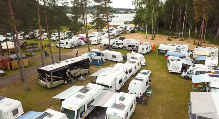 Duży zlot caravaningowy nad jeziorem. Wiele kamperów i przyczep kempingowych zaparkowanych na trawie, otoczonych drzewami.