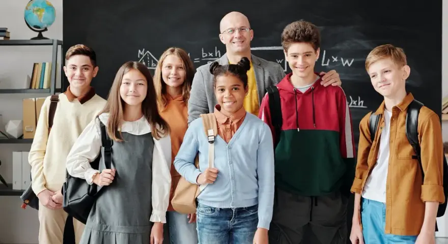 Uśmiechnięty nauczyciel i uczniowie przed tablicą. Czy szkoła społeczna to miejsce, gdzie tak dobrze się czują?