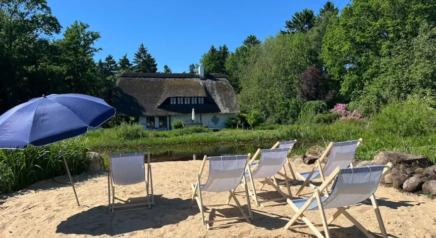 Sonnige Tage am Stedener See: Liegestühle und Sonnenschirm warten auf Besucher vor einem Reetdachhaus.