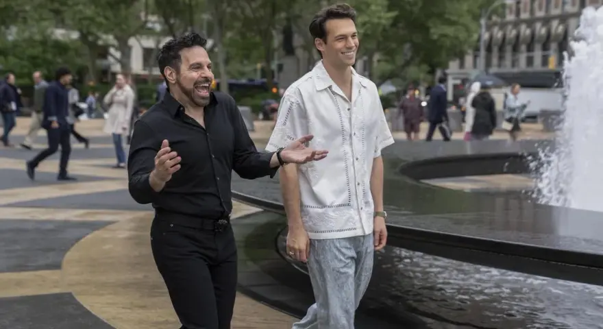 Il cast di And Just Like That, Mario Cantone e Billy Eichner passeggiano ridendo vicino a una fontana in una giornata nuvolosa.