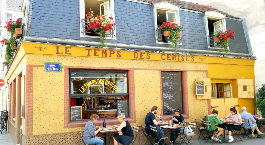 Café "Le Temps des Cerises" con un encantador estilo francés, balcones floridos y gente disfrutando en sus mesas exteriores.