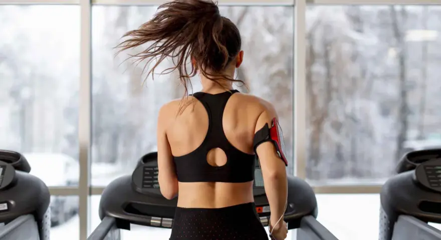 Une femme fait du sport à jeun sur un tapis roulant, ses cheveux flottant au vent. Dehors, la neige tombe.