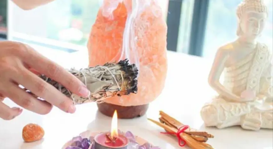 Manos sostienen un manojo de salvia humeante, junto a una lámpara de sal rosa, una vela encendida y una estatua de Buda. Esencial para la limpieza energética de casa.