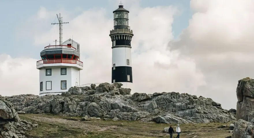Deux phares se dressent sur une côte rocheuse à Ouessant. Que faire ? Se promener et admirer le paysage, comme ces deux randonneurs.